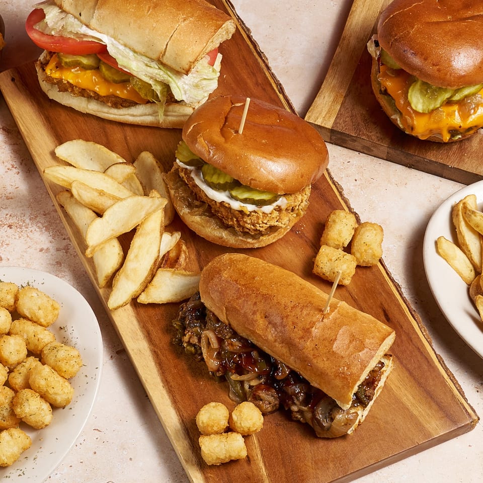Assorted cheeseburgers and sandwiches with pickles, lettuce, tomatoes, fries, and tater tots arranged on wooden serving boards.