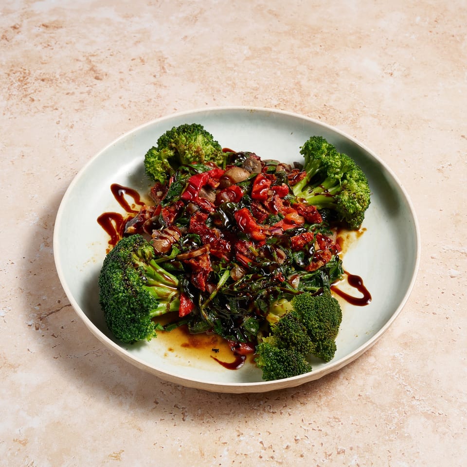 Plate of roasted broccoli, mushrooms, peppers, leafy greens, and sun-dried tomatoes drizzled with dark balsamic glaze.